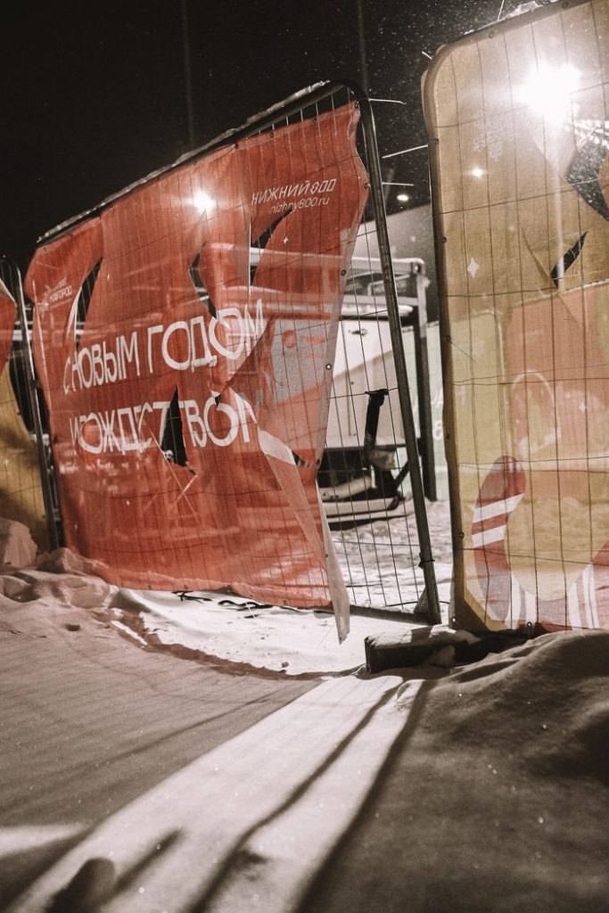 Snow-covered urban street scene at night with torn banner and fence.