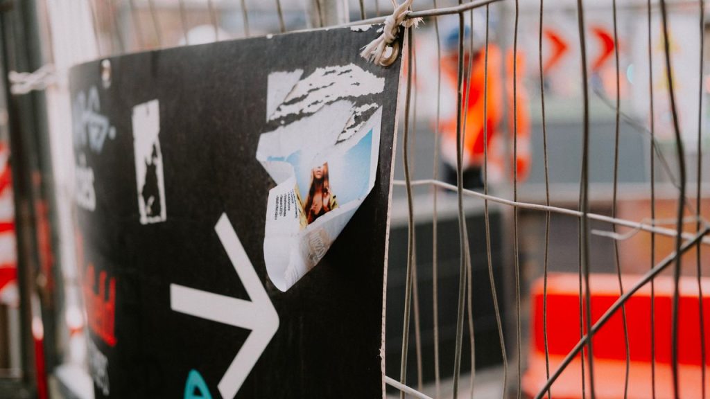 A torn poster featuring a person hangs on a metal fence, with a blurred background, outdoors.