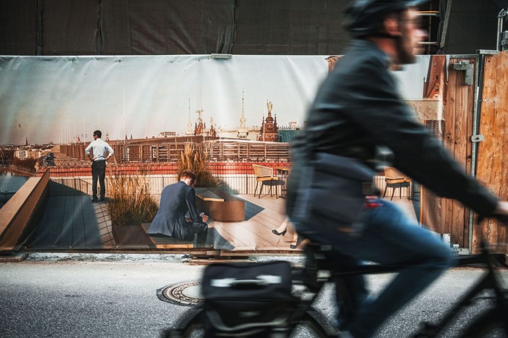 Bicycle rider passing by a construction site with a large photo poster depicting an urban rooftop scene.